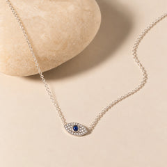 Silver evil eye necklace in 925 sterling silver with sparkling zircon accents and deep-blue center, styled on a soft ivory stone