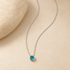 925 sterling silver blue zircon necklace with a petite round turquoise-blue pendant laid naturally beside a smooth beige stone on warm ivory background