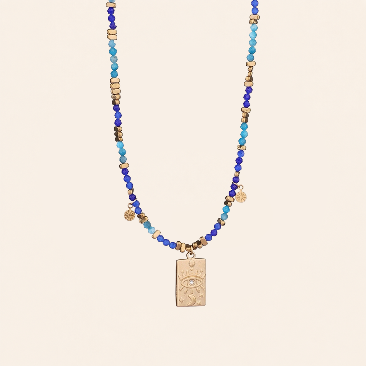 A Evil Eye pendant necklace made with natural blue stone beads featuring blue and turquoise beads, small metallic gold spacer beads, and a central gold-toned rectangular pendant with an eye design, displayed against a solid light beige background.
