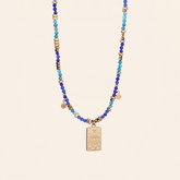 A Evil Eye pendant necklace made with natural blue stone beads featuring blue and turquoise beads, small metallic gold spacer beads, and a central gold-toned rectangular pendant with an eye design, displayed against a solid light beige background.