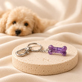 Purple natural crystal dog bone charm with a silver lobster clasp and keyring, displayed on an ivory stone pedestal with a softly blurred puppy in the background.