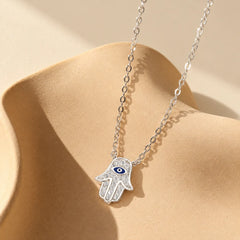 Silver Hamsa evil eye necklace with pavé-style zircon details on an ivory sculptural ceramic display