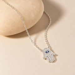 Silver Hamsa hand necklace with zircon-set blue evil eye pendant draped across a rounded ivory stone
