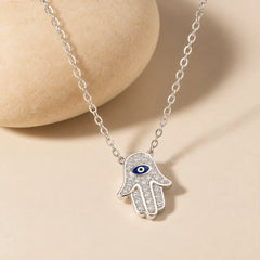 925 sterling silver Hamsa hand necklace with blue evil eye and zircon-set pendant beside a smooth warm ivory stone