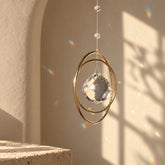 Golden Orbit clear quartz crystal suncatcher hanging in warm sunlight with rainbow reflections on a neutral wall