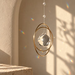Golden Orbit clear quartz crystal suncatcher hanging in warm sunlight with rainbow reflections on a neutral wall