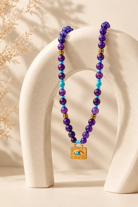 Amethyst Evil Eye necklace with purple crystal beads, turquoise accents, and a gold protective eye pendant on a cream stone arch