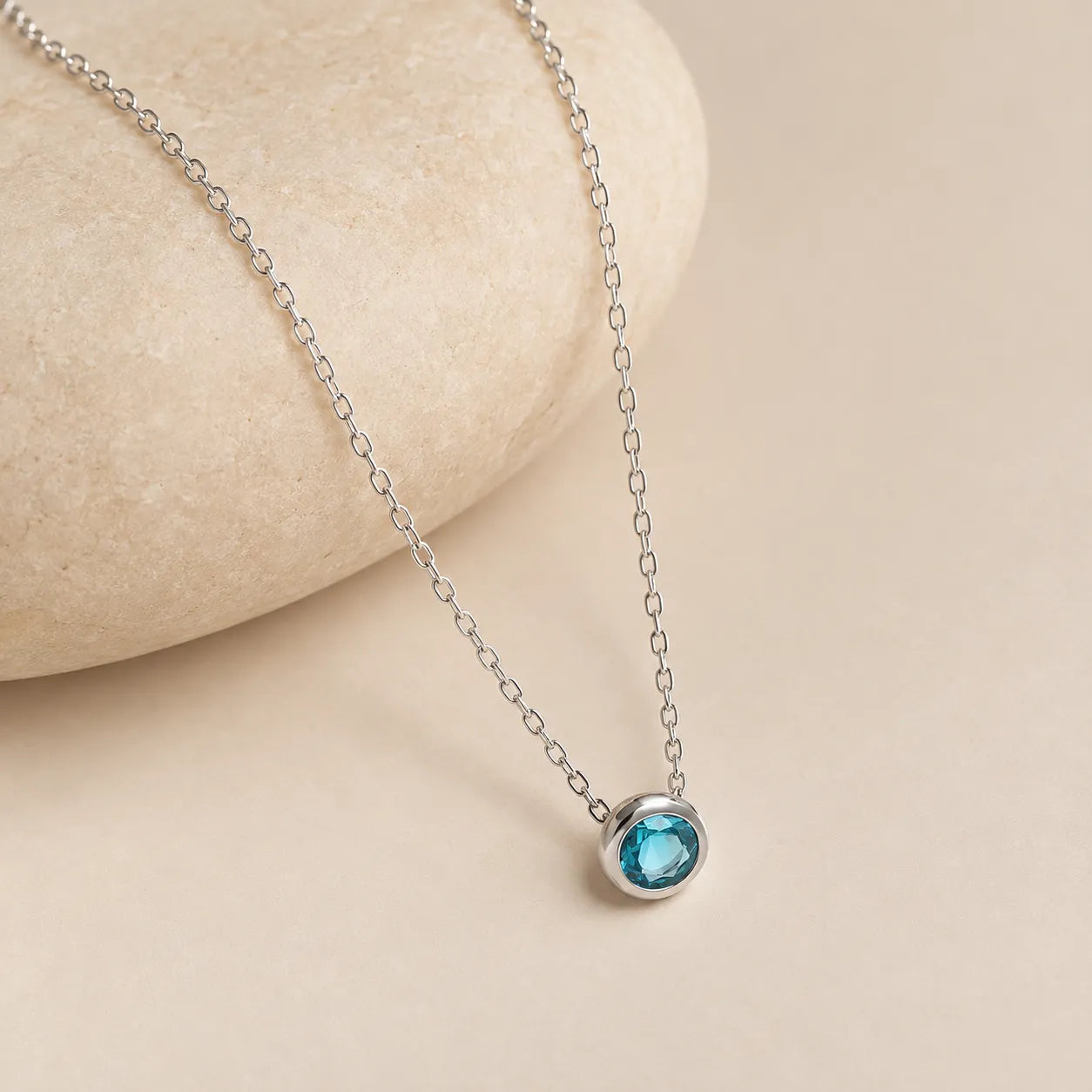 Dainty blue zircon necklace in sterling silver with small round bezel-set gemstone and naturally hanging chain beside a soft beige stone