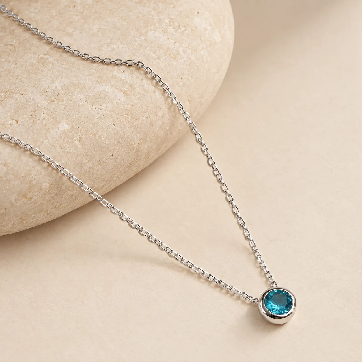 Close-up of a faceted blue-green zircon pendant in a polished silver bezel, with fine cable chain draped over a rounded ivory stone
