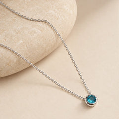 Close-up of a faceted blue-green zircon pendant in a polished silver bezel, with fine cable chain draped over a rounded ivory stone