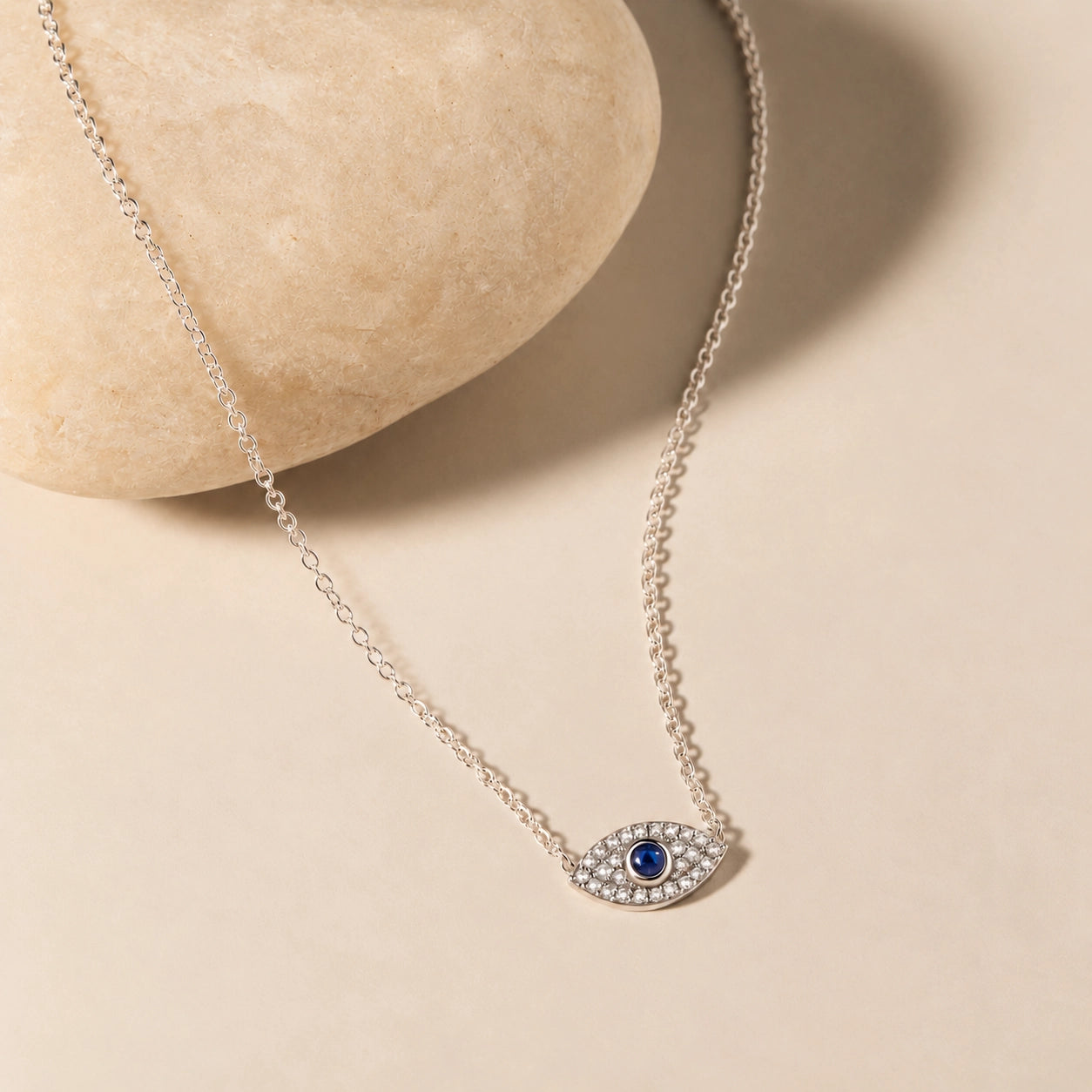 Delicate sterling silver evil eye necklace with a zircon-set blue eye pendant styled against a natural warm ivory stone