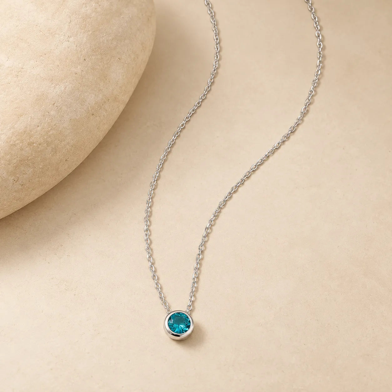 925 sterling silver blue zircon necklace with a petite round turquoise-blue pendant laid naturally beside a smooth beige stone on warm ivory background