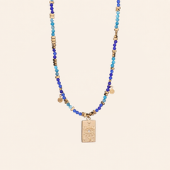 A Evil Eye pendant necklace made with natural blue stone beads  featuring blue and turquoise beads, small metallic gold spacer beads, and a central gold-toned rectangular pendant with an eye design, displayed against a solid light beige background.