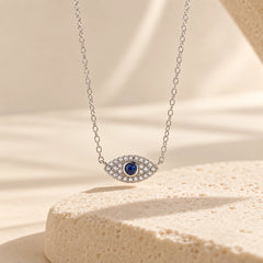 Silver sterling silver evil eye necklace with blue center stone and zircon accents displayed above a textured ivory pedestal