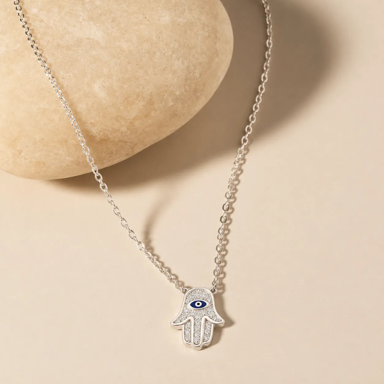 Dainty sterling silver Hamsa hand necklace with sparkling zircon and blue evil eye pendant on soft ivory surface