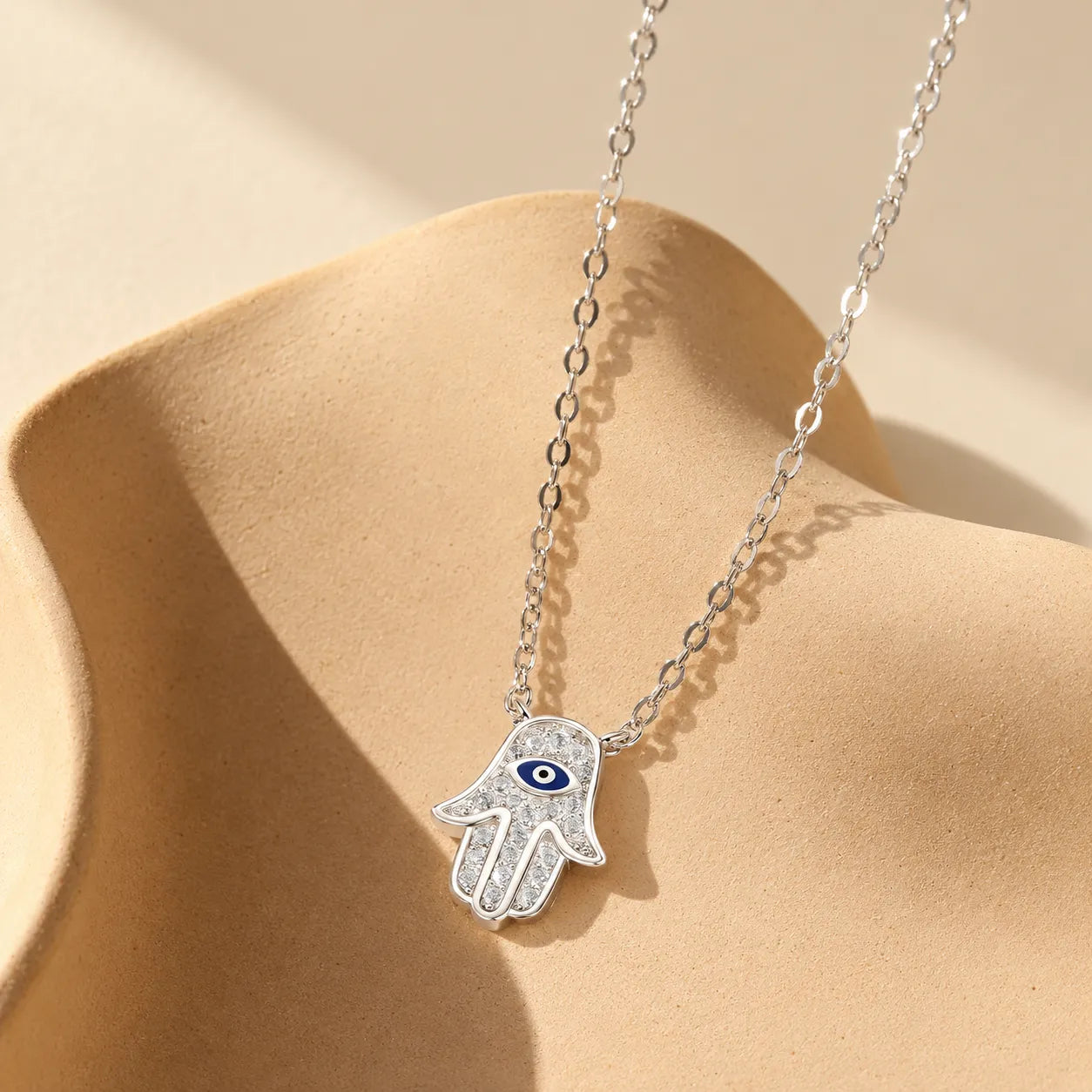 Silver Hamsa evil eye necklace with pavé-style zircon details on an ivory sculptural ceramic display