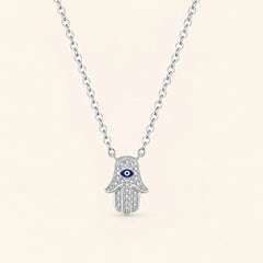 Front view of sterling silver Hamsa hand necklace featuring a blue evil eye motif and shimmering zircon stones on ivory background