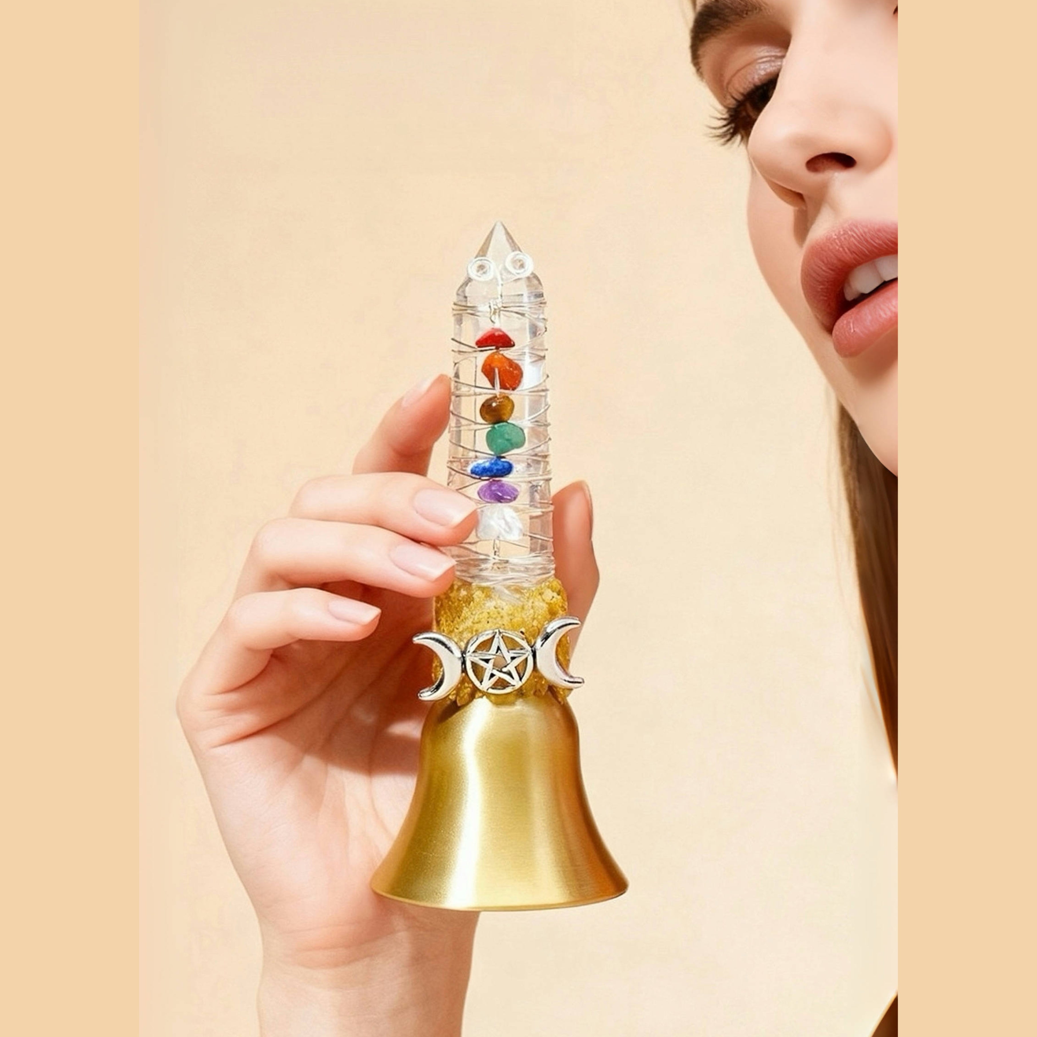 Hand holding a crystal point bell with chakra colors and a gold bell against a beige background