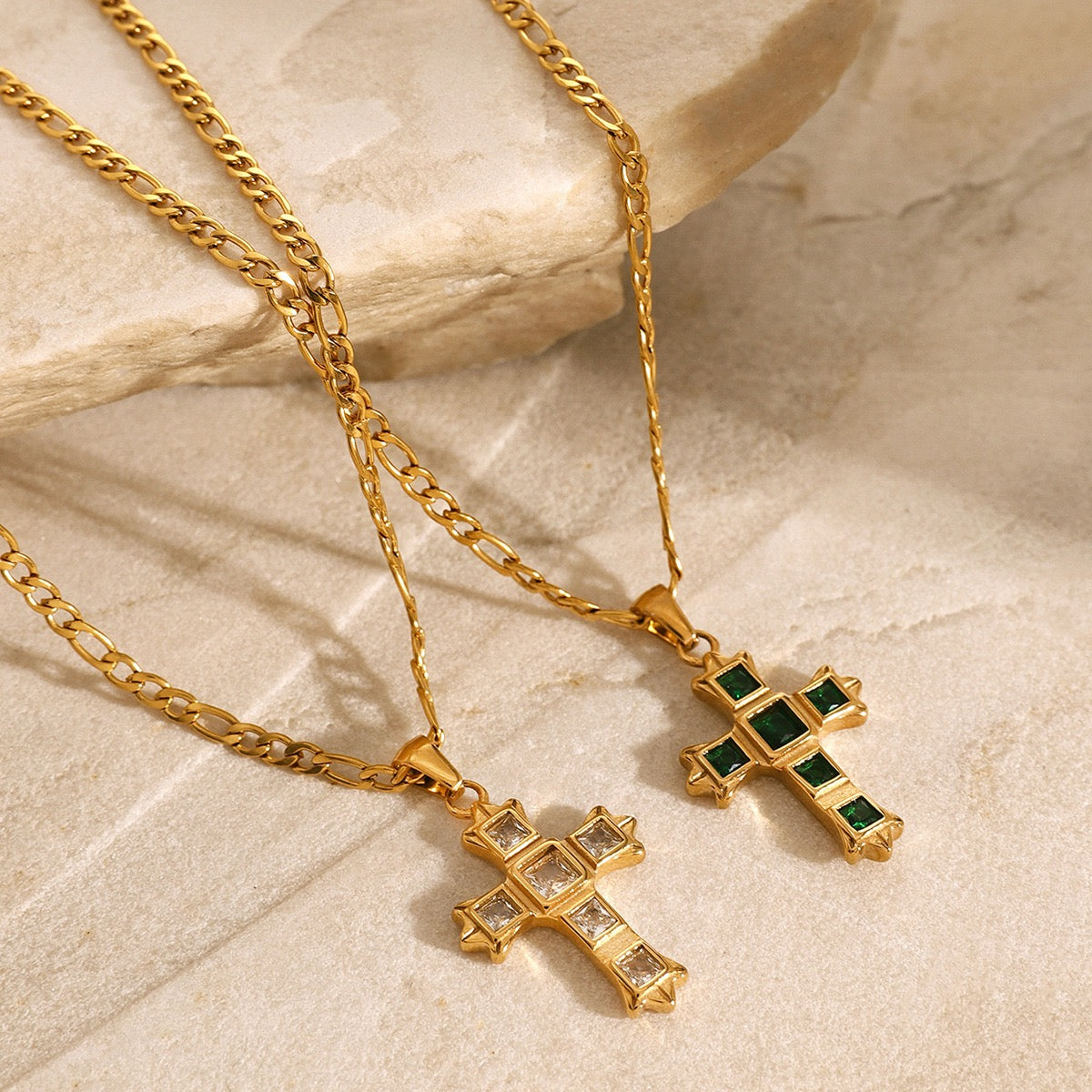 Two gold cross necklaces with gemstone accents on a beige stone surface
