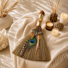 Handmade peacock feather witch broom displayed on soft beige fabric with candles and cozy home decor