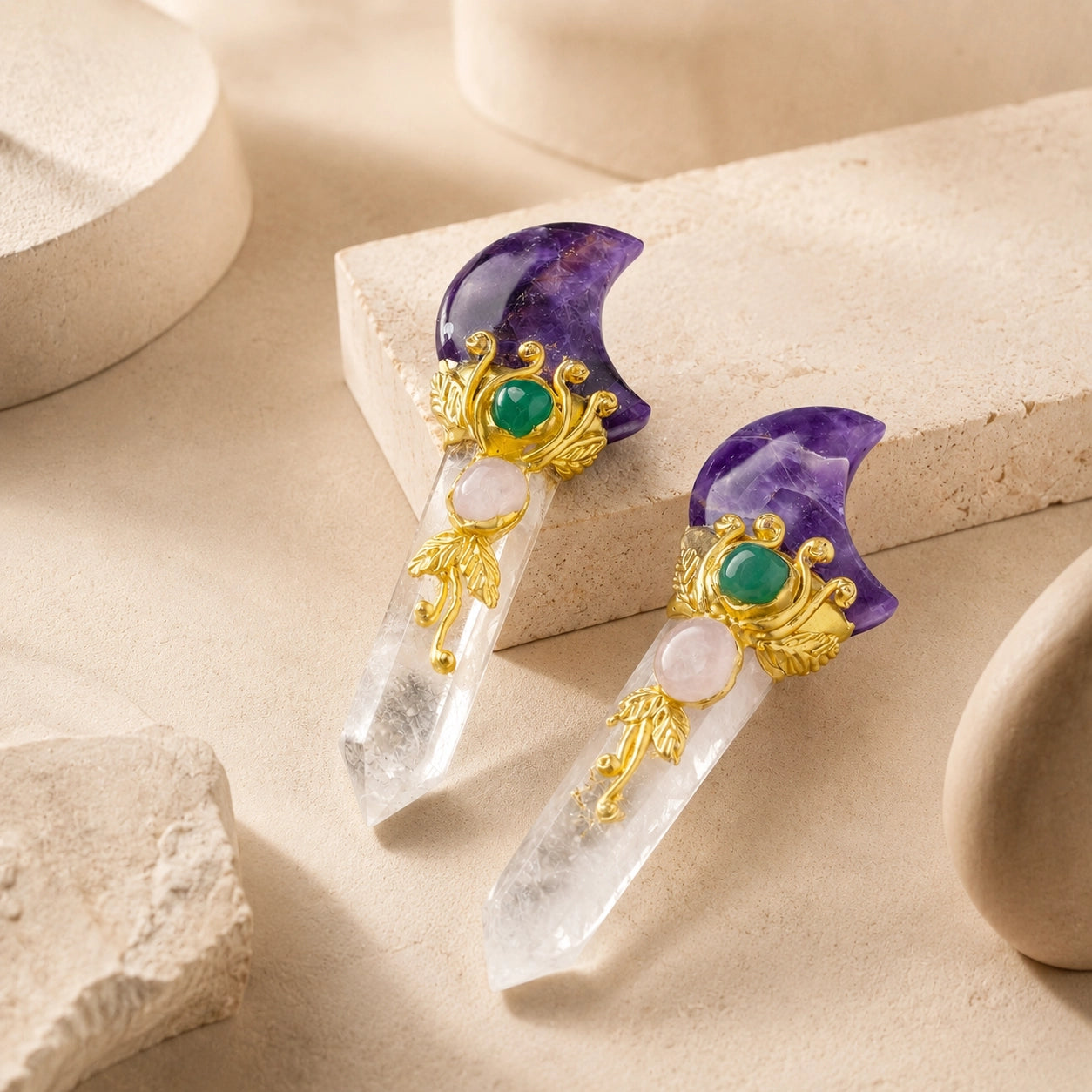 Two Amethyst crescent moon crystal point wands with gold detailing lying flat on warm, natural beige display stones.