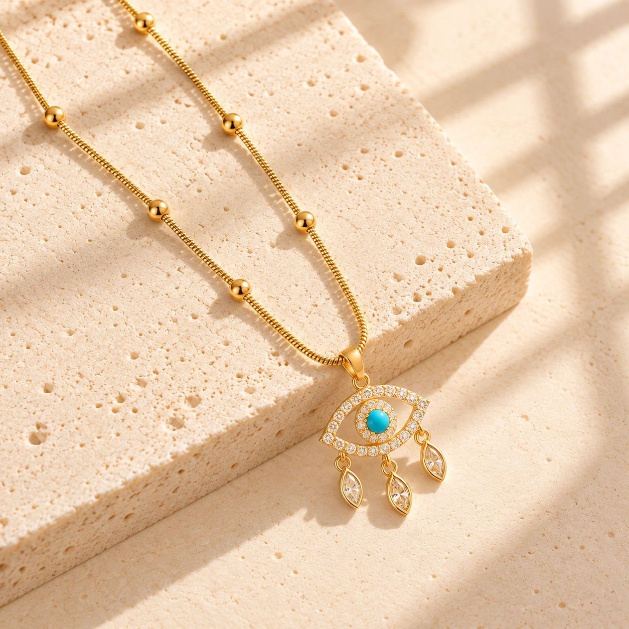 Blue evil eye pendant necklace with gold-tone beaded chain laid on warm ivory travertine
