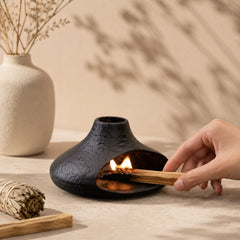 Hand placing a lit Palo Santo stick inside a black chimney-style ceramic burner