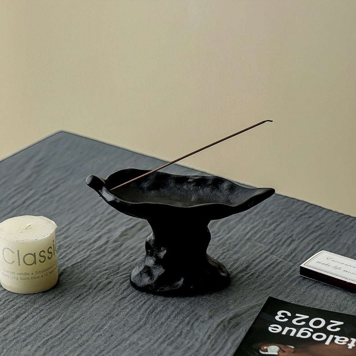 Black incense burner with a burning stick on a textured surface next to a candle and a book.